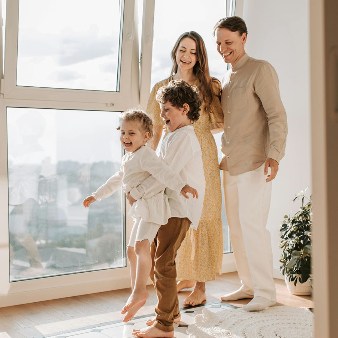 A family of four, two adults and two children, are standing by a large window